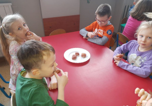 Dzieci przy stoliczku czerwonym z wielkim apetytem zjadają pączki.