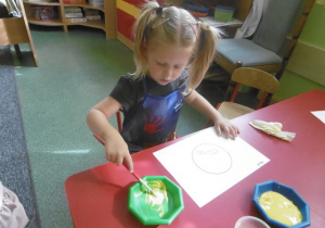 Nela maluje farbami słoneczko - symbol przedszkola.