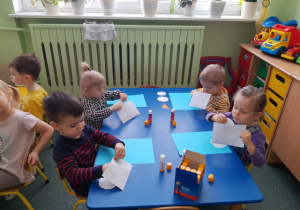 Dzieci siedzące przy stoliku niebieskim wyjmują gotowe elementy pracy plastycznej z koperty.