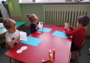 Dzieci siedzące przy stoliku czerwonym wyjmują gotowe elementy pracy plastycznej z koperty.