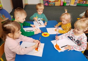 Dzieci przy stoliku niebieskim malują parasole pomarańczową farbą