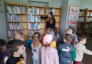 Pani bibliotekarka pokazuje dzieciom regał z ciekawymi książkai.