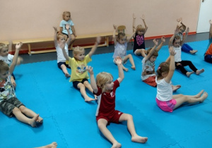 Dzieci ćwiczą w pozycji siedzącej z podniesionymi rękami .