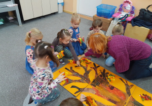 Dzieci przyklejają listki do pomalowanego drzewa.
